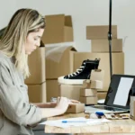 Seorang perempuan sedang mengemas paket di depan laptop untuk bisnis online, menggambarkan aktivitas reseller atau dropshipper